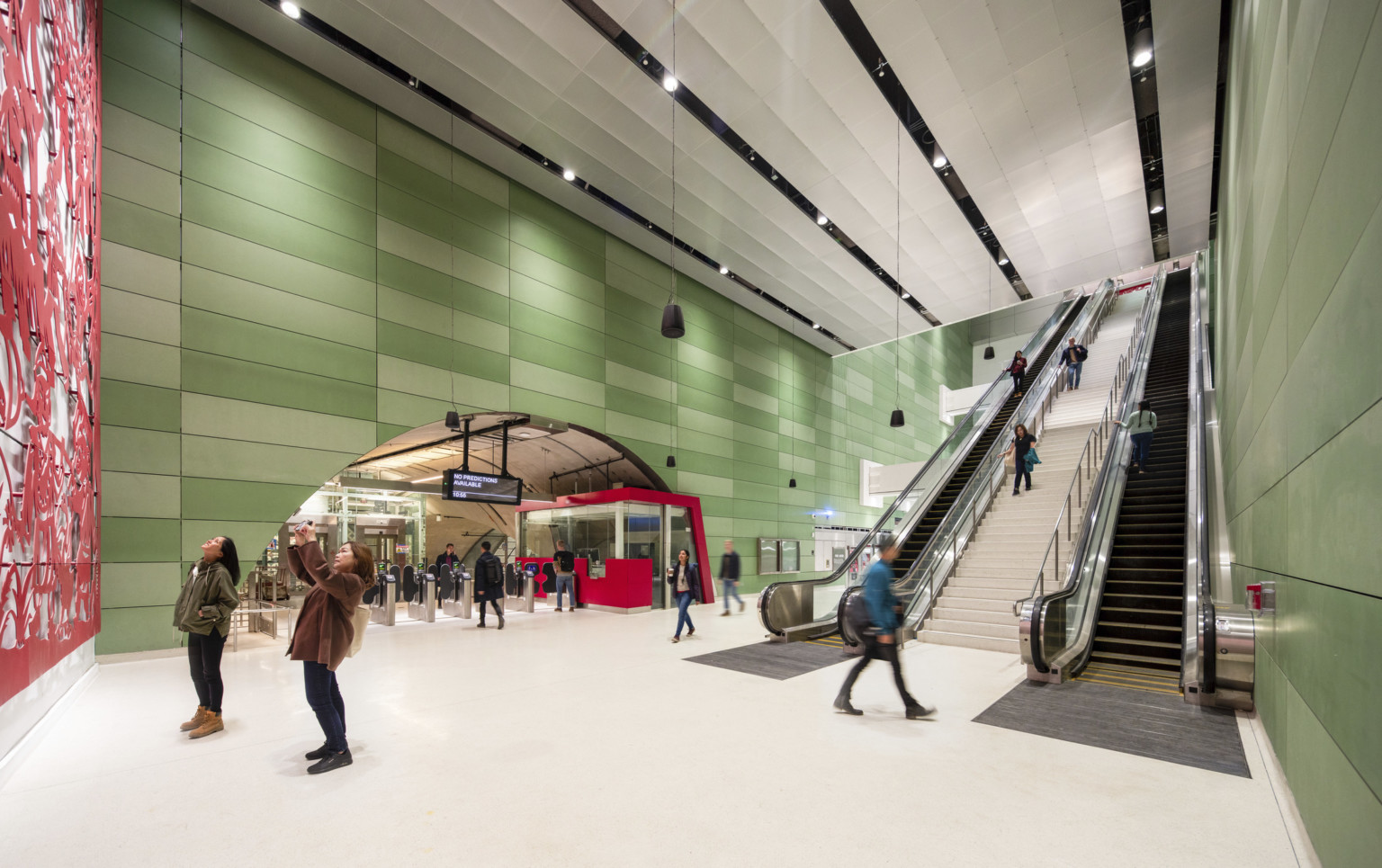 2 people looking at red art mural in a sub ground train station, escalators to back, rounded entry over turnstiles left