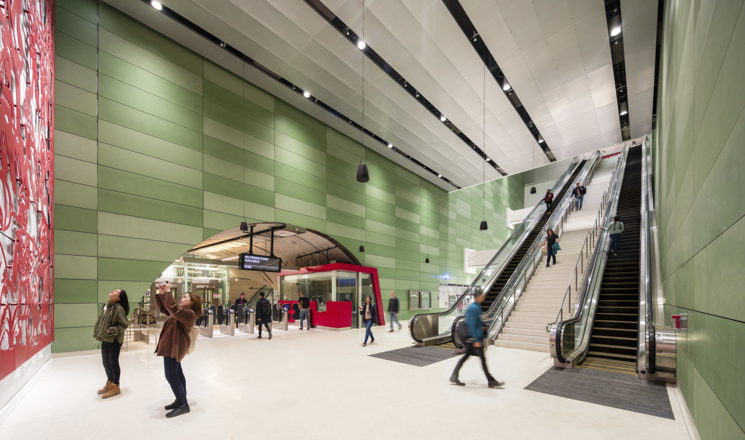 2 people looking at red art mural in a sub ground train station, escalators to back, rounded entry over turnstiles left