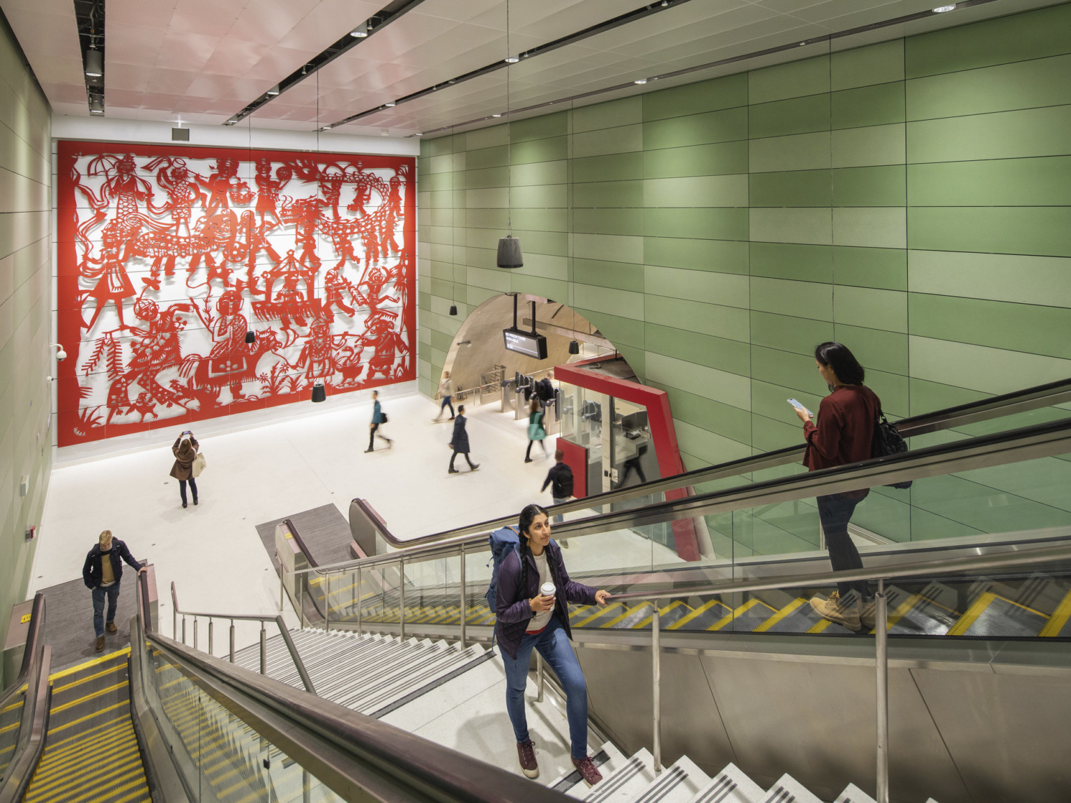 Concrete block walls with raised red sculptural mural on left. Right, arched hall with gates and ticket booth by escalators