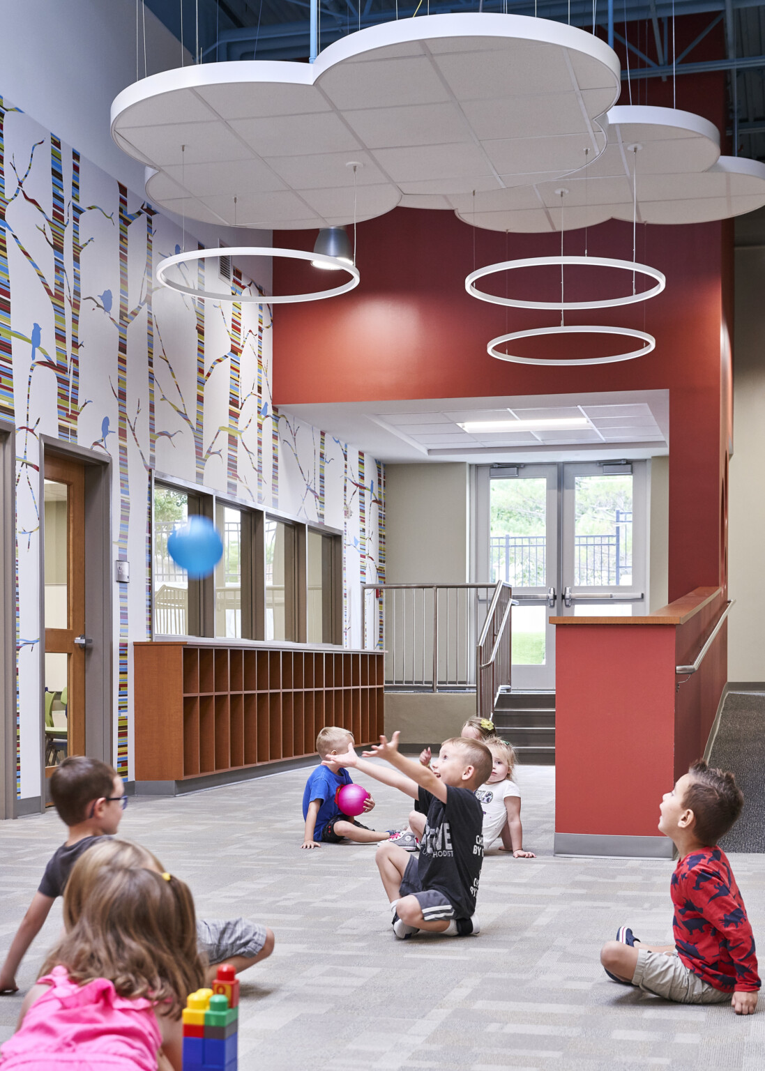 Red hallway with colorful striped tree mural on white wall, left. Hanging cloud details and circular lights over children playing on carpet