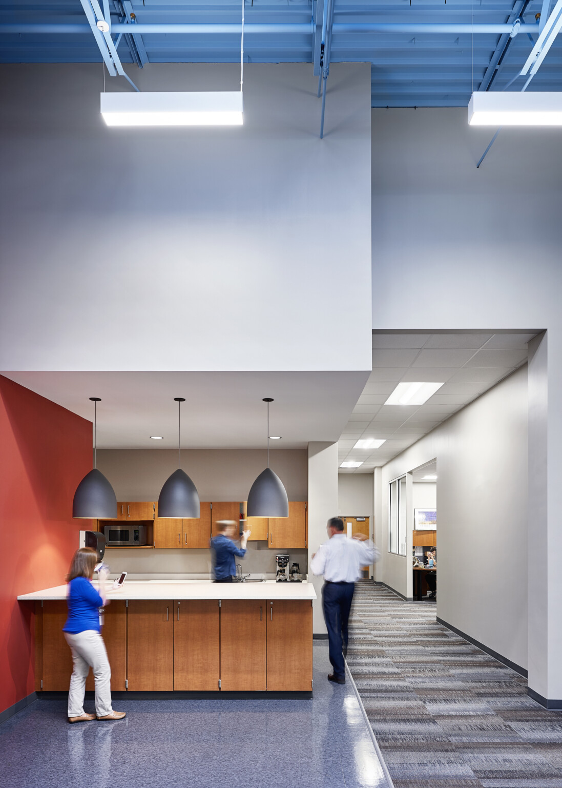 Kitchenette alcove in double height hallway with red accent wall and large pendant lights over island with wood cabinets