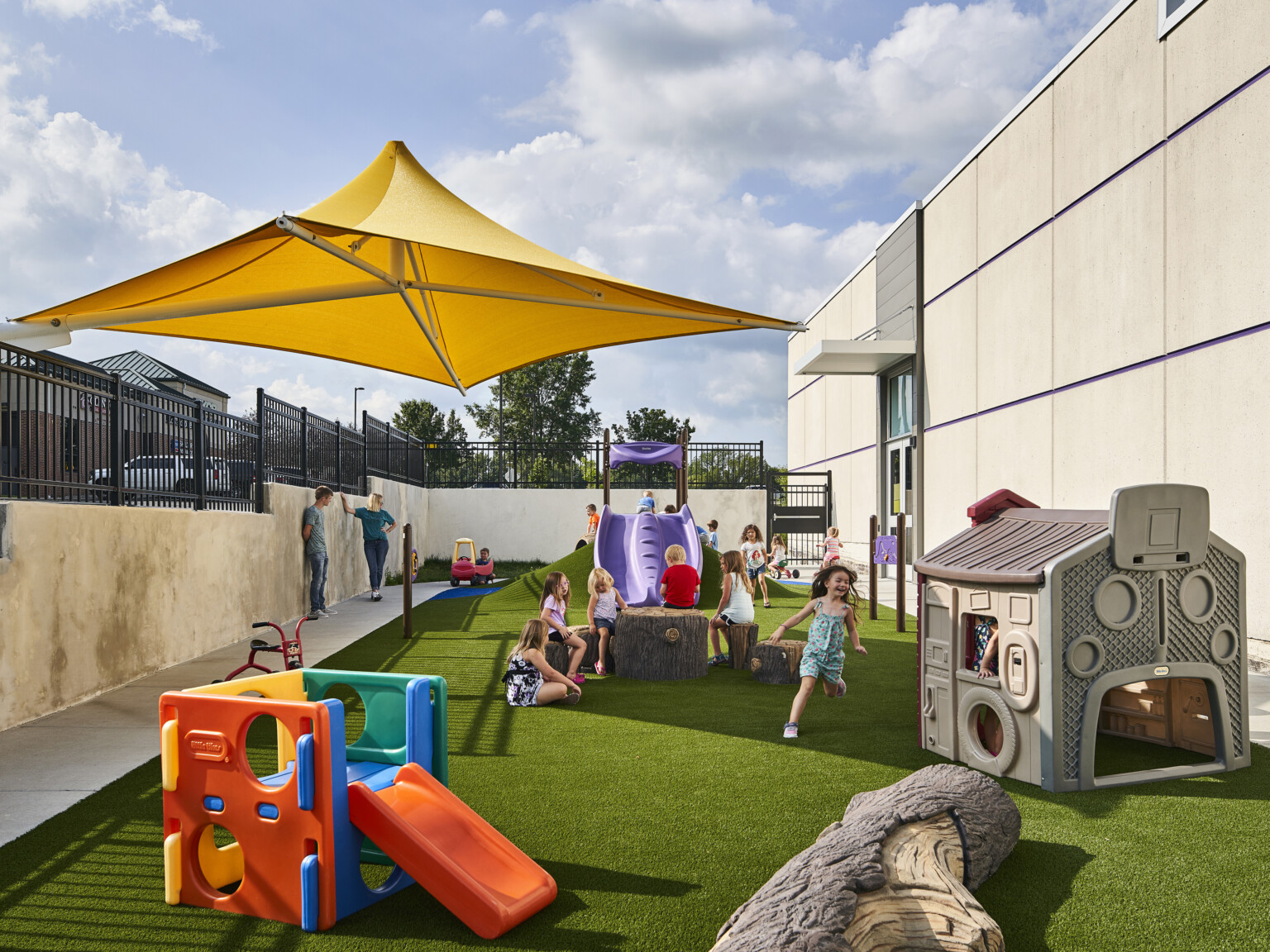 Playground with turf outside concrete building with yellow area umbrella partially covering. Man made hill with purple slide