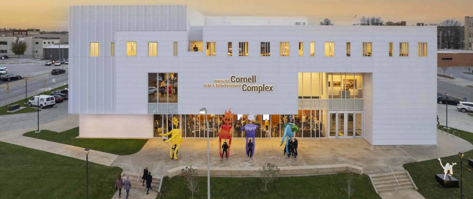 White multistory building light up at dusk by yellow lights; sculptures fill the lawn with people filing in to watch a show at the theater