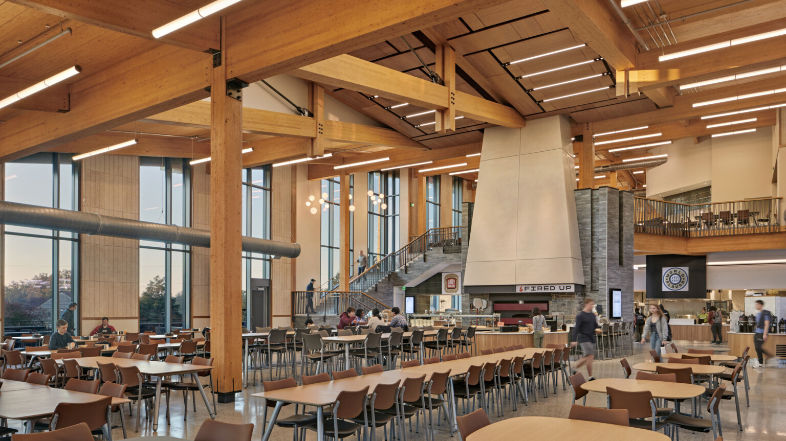 large two-story glass dining hall with exposed timber beams and posts, stone wrapped stair leading to second floor seating area
