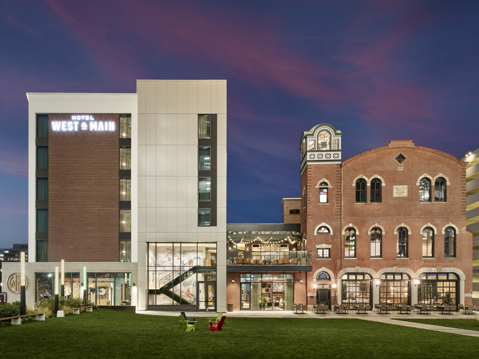 On the left, a new multi-story hotel wrapped in light siding with brick accents and on the right, the former brick fire station connects to the new hotel by an enclosed structure with a rooftop balcony overlooking a lawn.