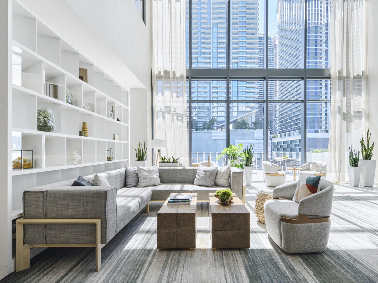 Bespoke white divider shelf in large group with gray, modern seating, wood block coffee table, and planters in double height glass room