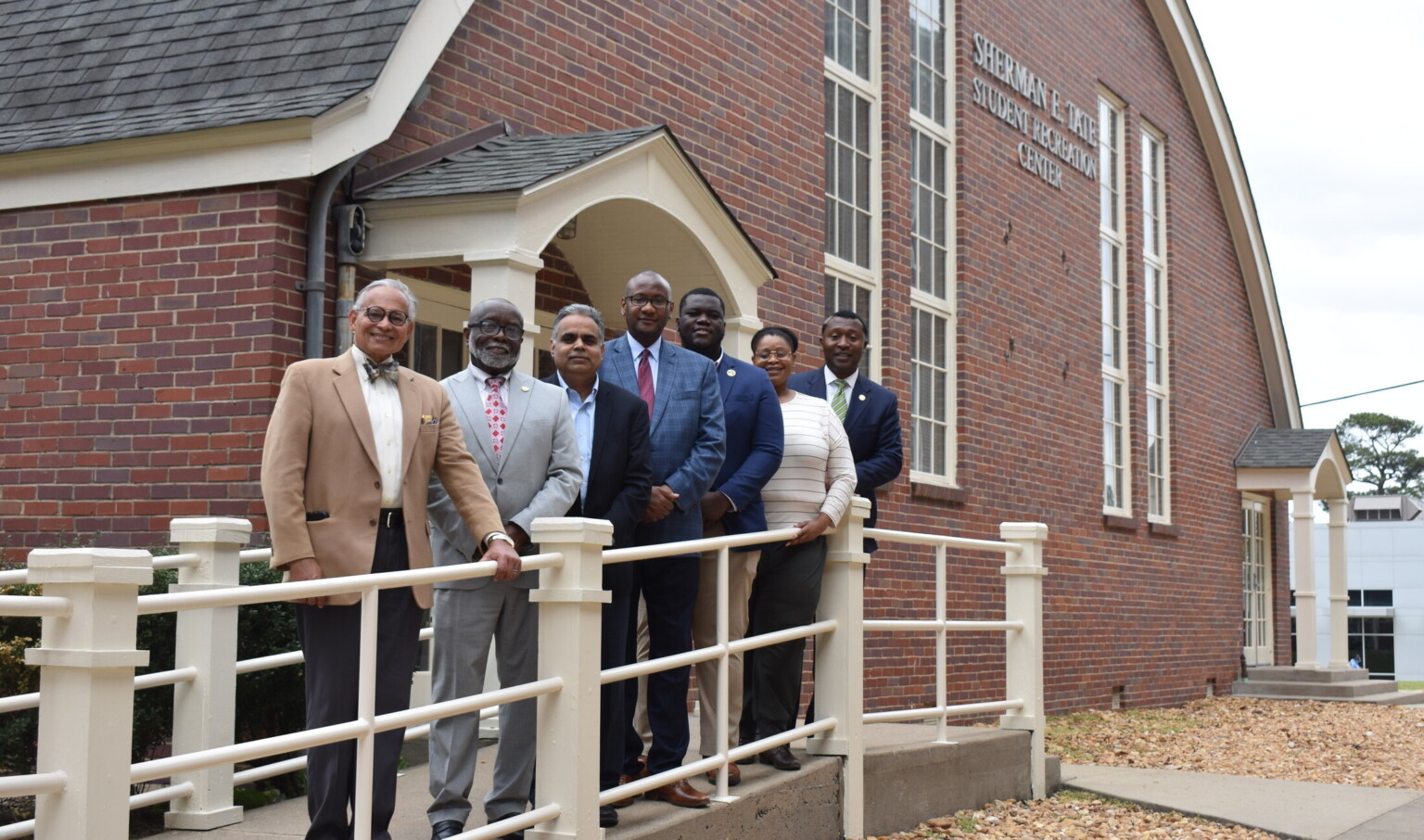 people in suits pose for photo outside a red brick building, arched roof and windows on a ramp with white metal railing,