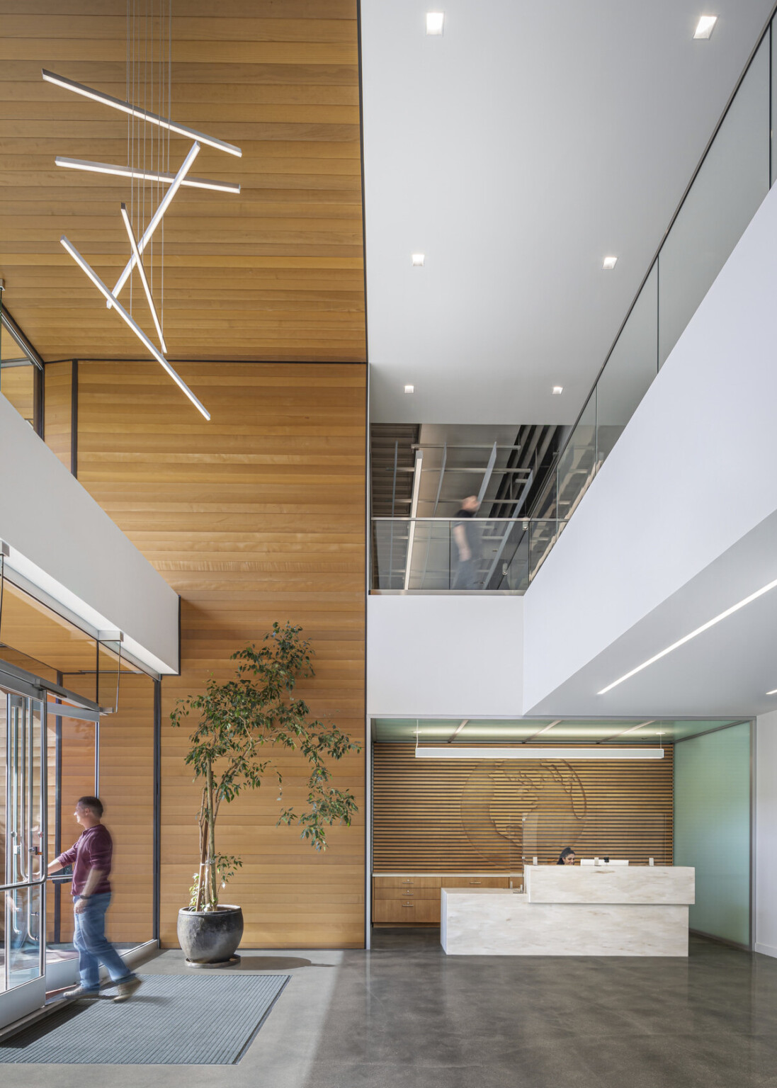 Double height entry lobby with wood panel and white balcony with glass rail. Large windows and hanging sculpture