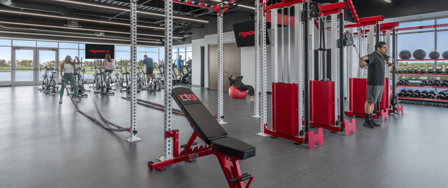 Fitness center with strength equipment, stationary bikes. Industrial ceiling with exposed ducts. Floor to ceiling windows, lake view