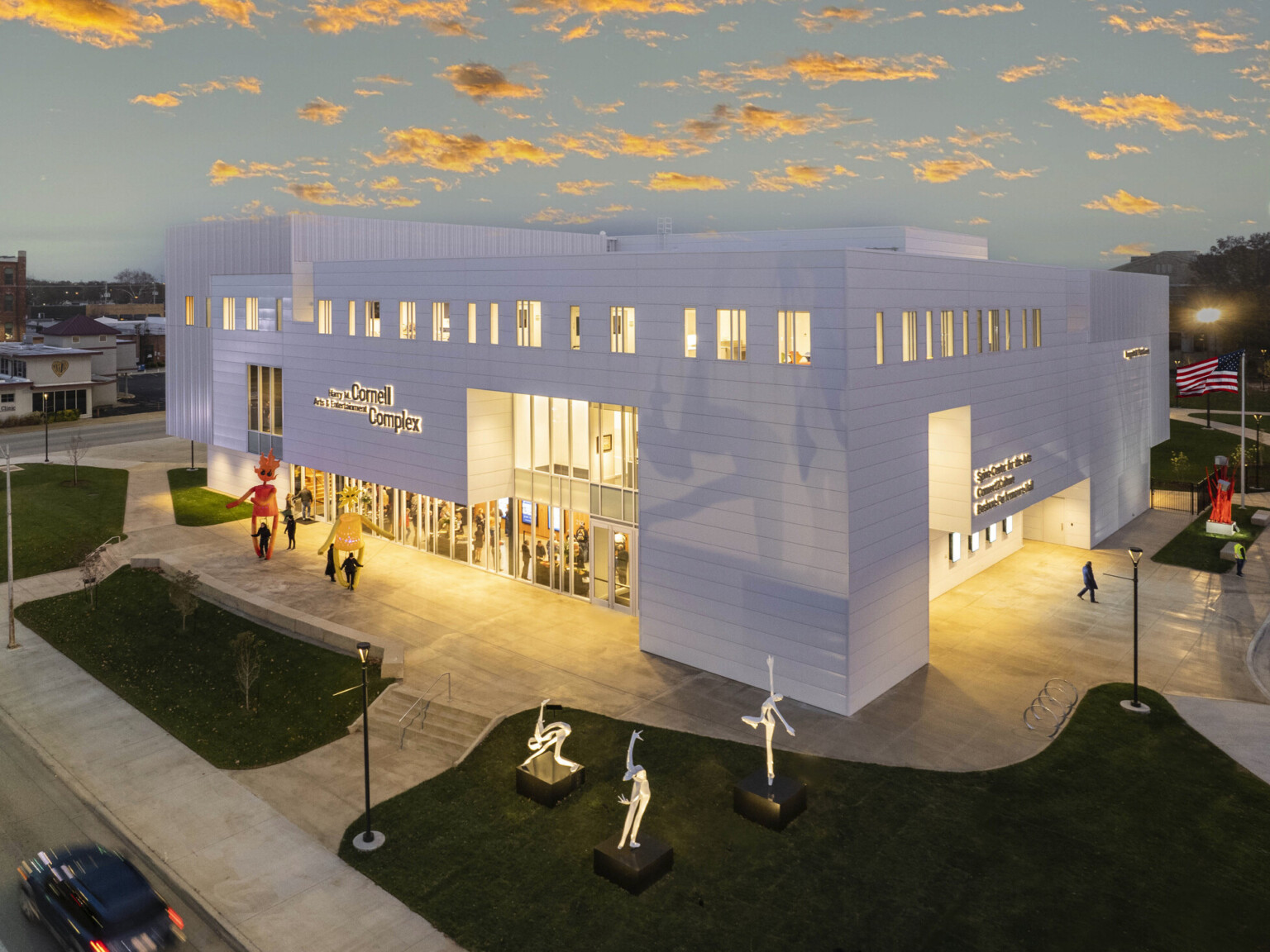 Exterior, night-time view of the new Harry M. Cornell Arts & Entertainment Complex, a white paneled building, sculptures on lawn