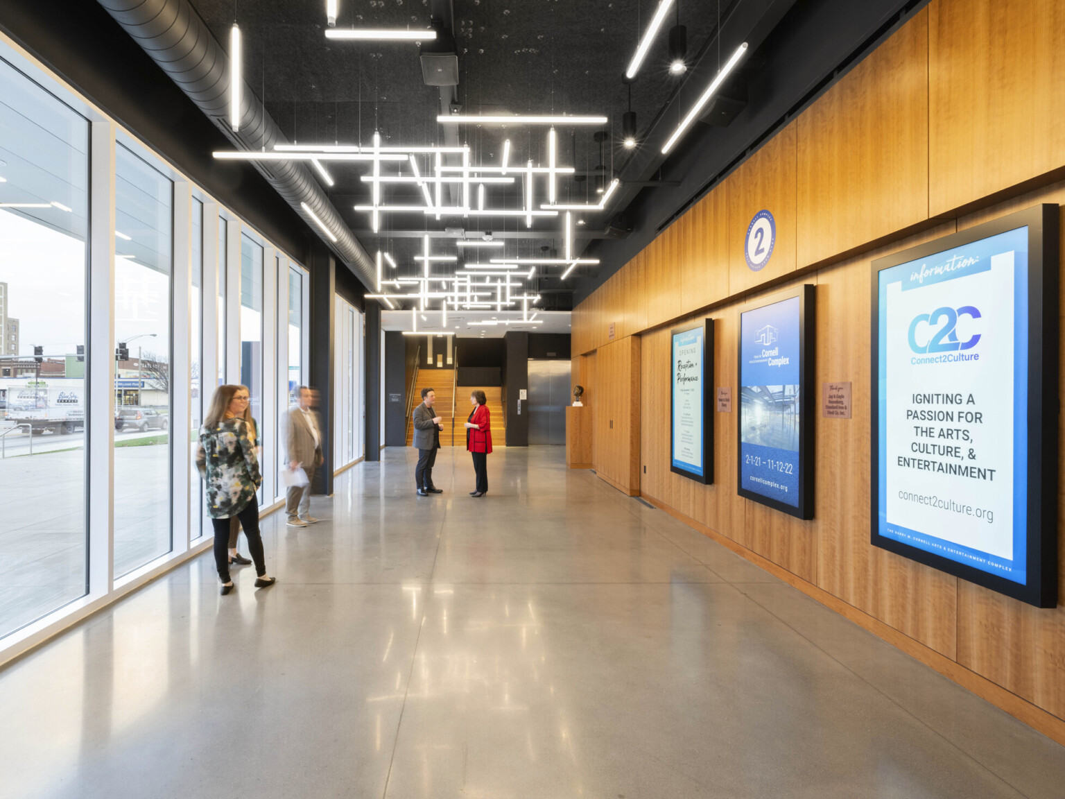 Brightly lit hallway, floor to ceiling windows, glass wall to left. Wood panel wall with posters, right. Sculptural pendant lights