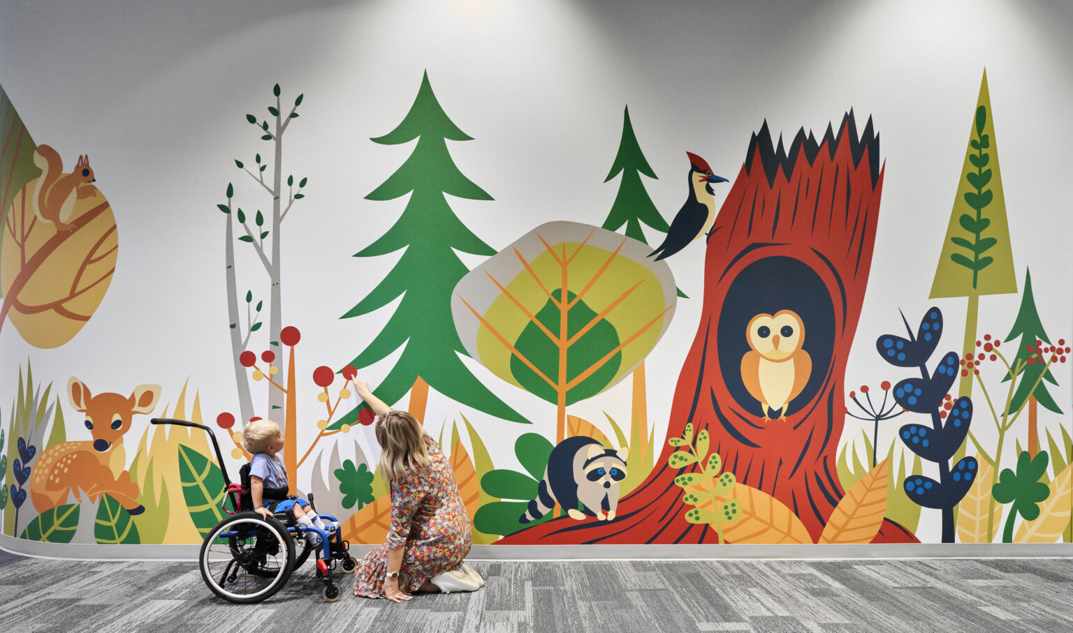 Child and educator interact with mural of cartoon forest and animals in carpeted school hallway. Mixed trees, deer, owl, racoon