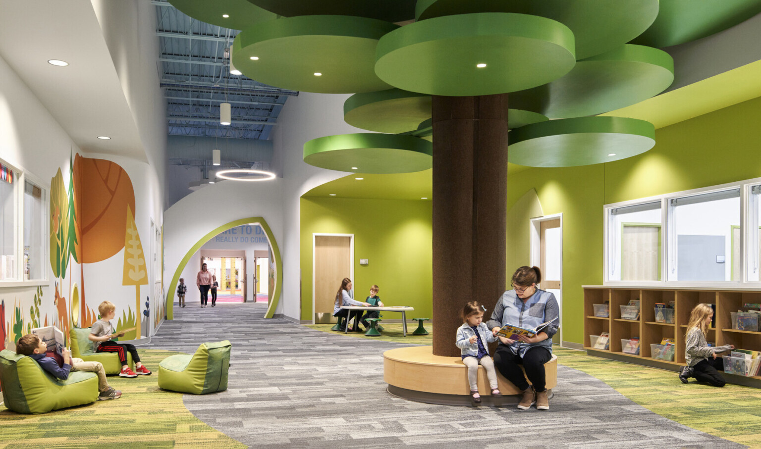 people reading on a brown circular bench with a brown column and green ceiling fixtures in hallway lined with forest graphics