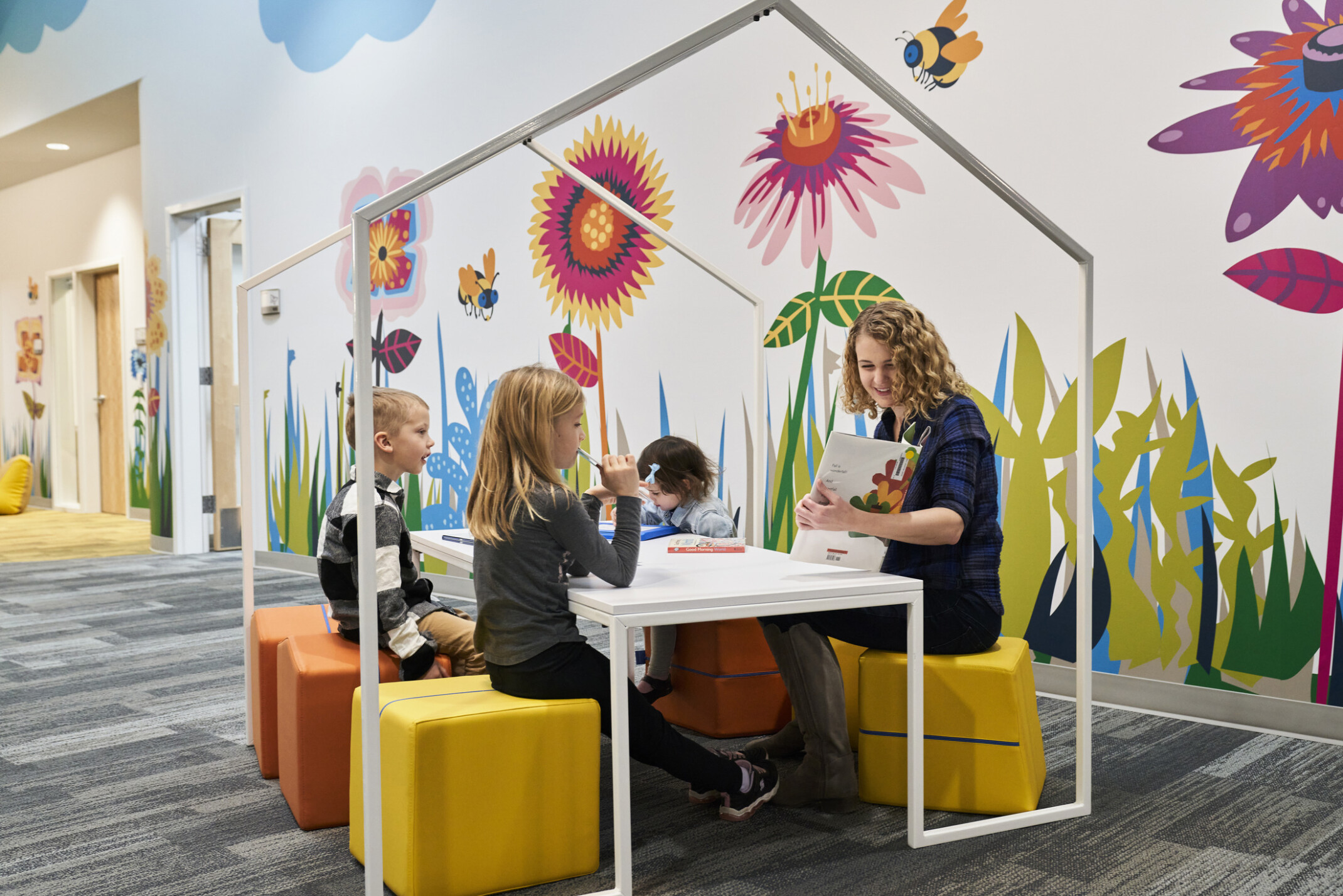 Educator and students reading on flexible comfortable yellow stools at white table with house shaped frame, bright graphic mural