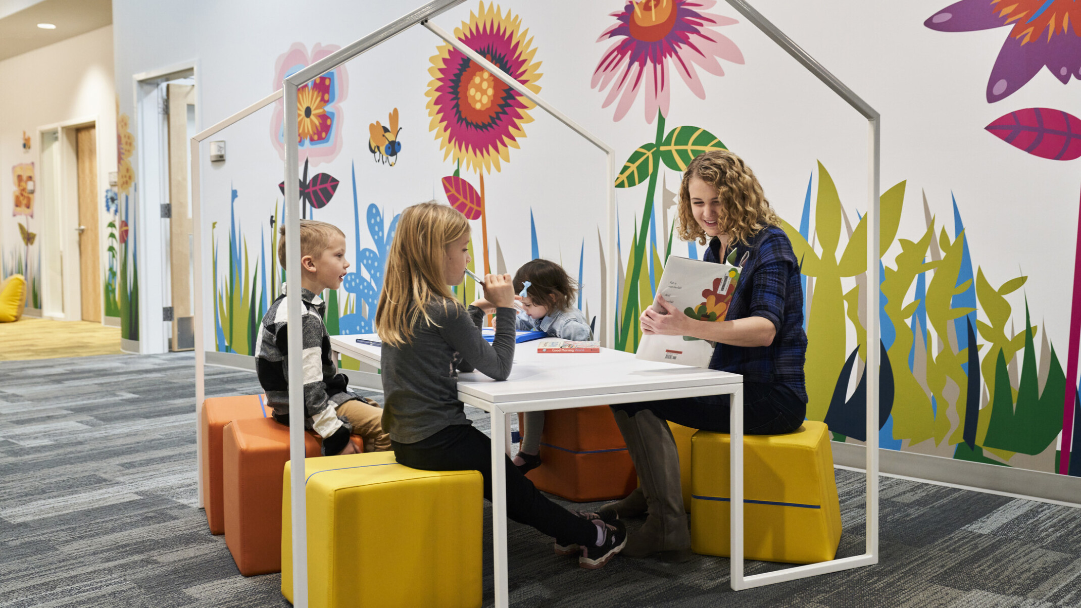 Educator and students reading on flexible comfortable yellow stools at white table with house shaped frame. Bright graphic mural