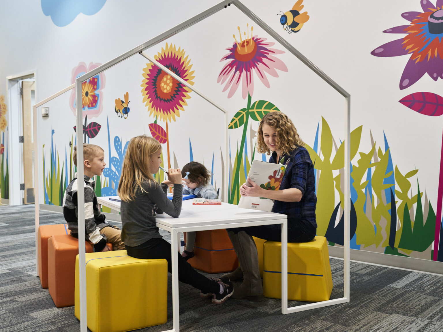 Educator and students reading on flexible comfortable yellow stools at white table with house shaped frame. Bright graphic mural