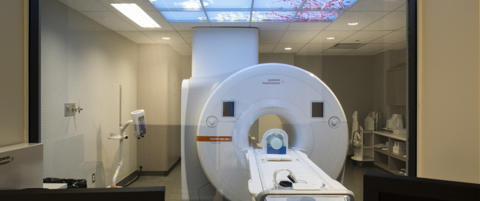 View through interior window to CAT Scan machine in white room with fake skylight showing blue sky with white fluffy clouds