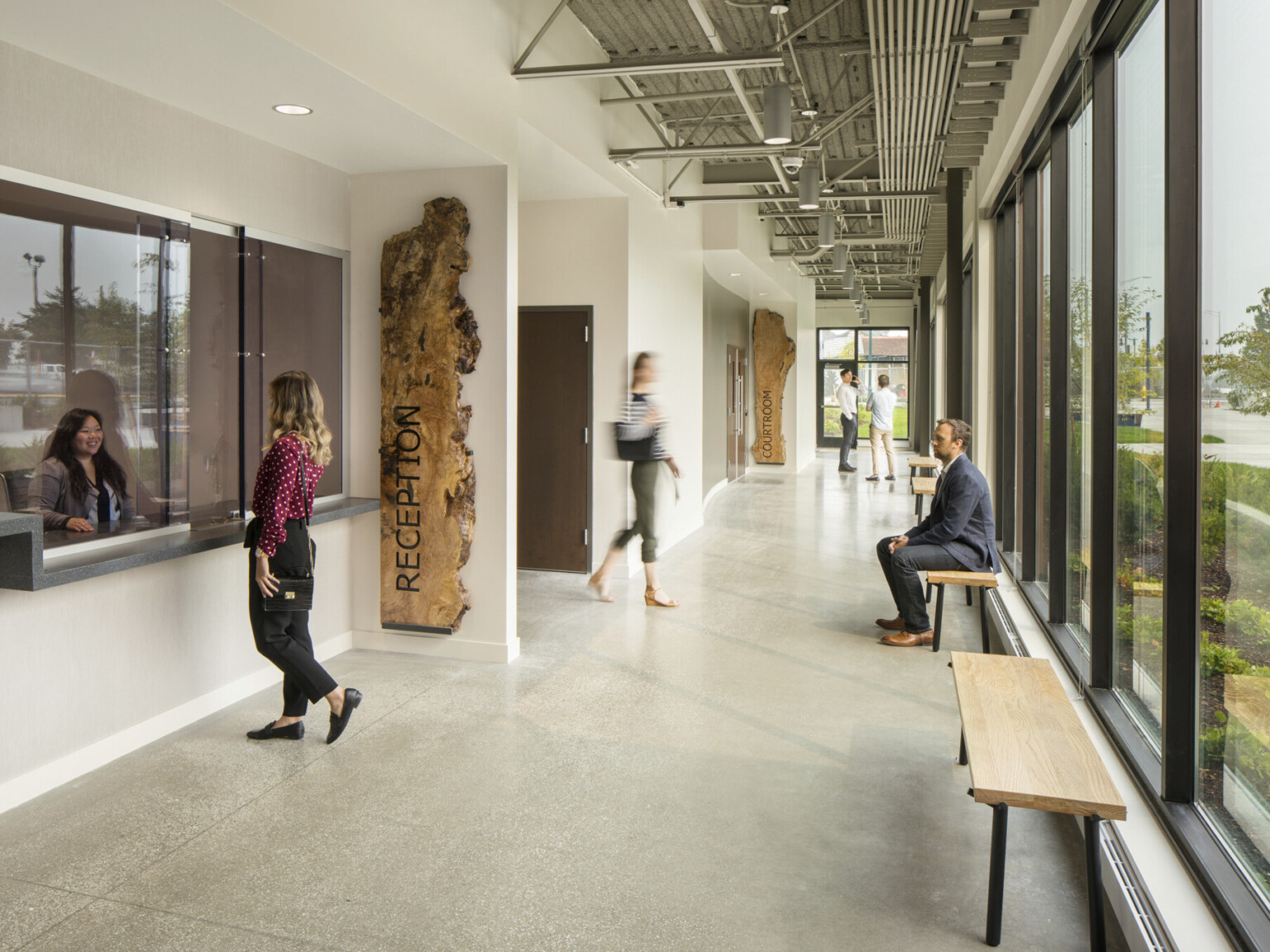 White hallway with tall ceilings and wall of windows. Reception desk, large wood organically shaped signage. Industrial ceiling