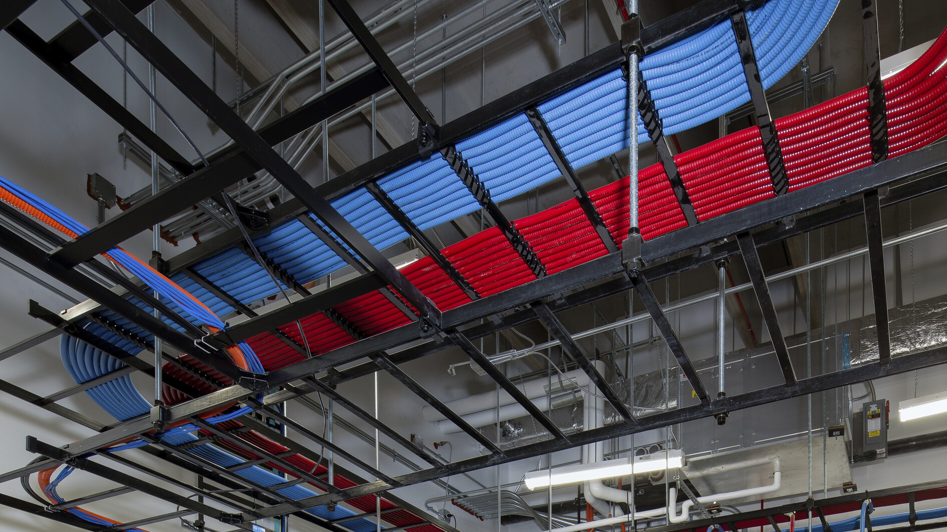 an open ceiling with black orthogonal framing system that contains lighting pendants, pipes, metallic and red and blue tubes