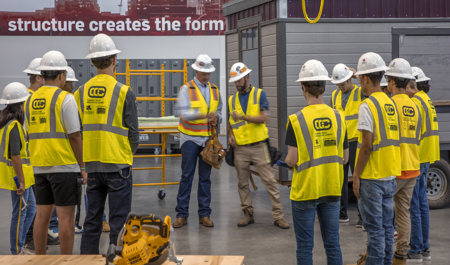 Circular saw on table in front of students in safety vest and hard hats circled around teachers. Paneled room, back right Circular saw on table in front of students in safety vest and hard hats circled around teachers. Paneled room, back right