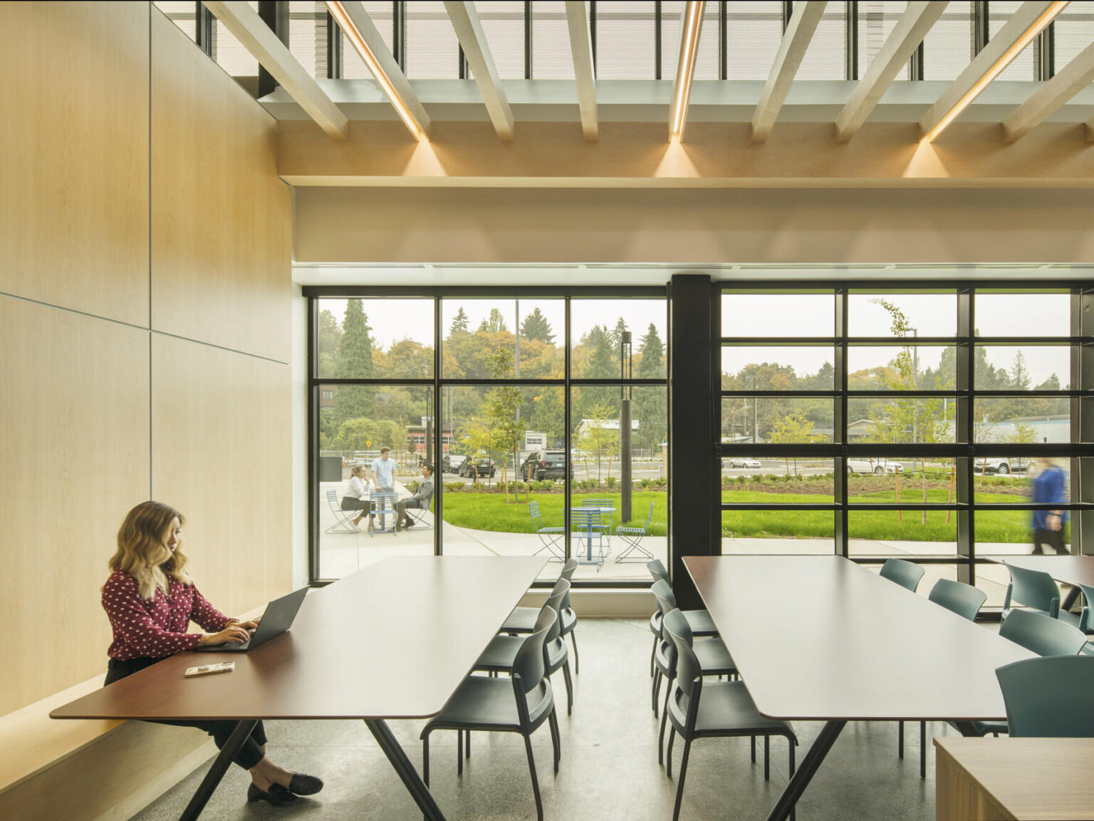 Interior seating area with chairs along long tables. Floor to ceiling windows look out onto grass courtyard. Slat ceiling canopy