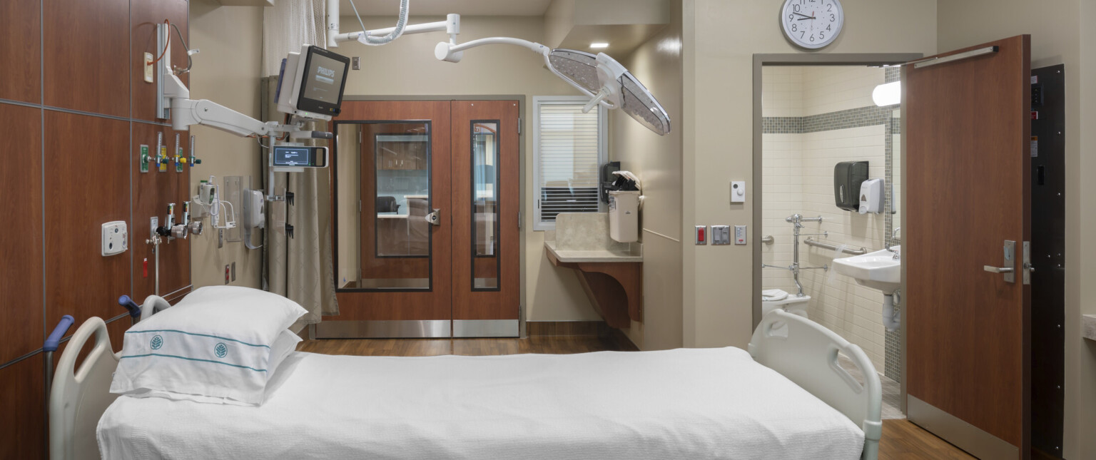 Hospital bed with equipment hanging overhead, brown wood accents on walls and doors of beige room with private bathroom