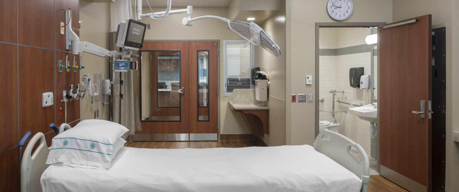 Hospital bed with equipment hanging overhead, brown wood accents on walls and doors of beige room with private bathroom Hospital bed with equipment hanging overhead, brown wood accents on walls and doors of beige room with private bathroom