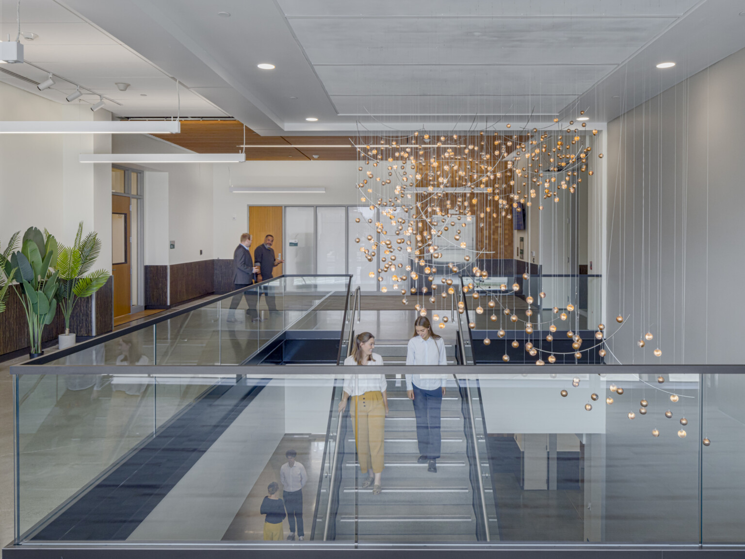 Warm wood tones and metal and glass staircase, high-ceilings, recessed lighting, modern chandelier, two women talking, walking down communicating staircase