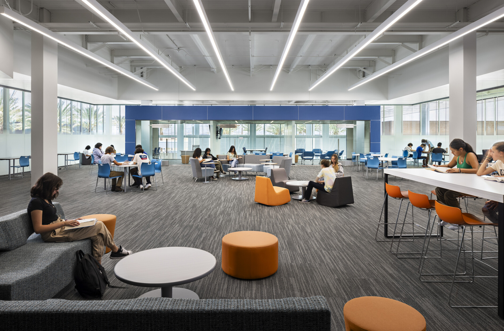Mixed comfortable seating; liberty high school common area; walls of windows surrounding room, high school design with white ceiling, blue and orange accents