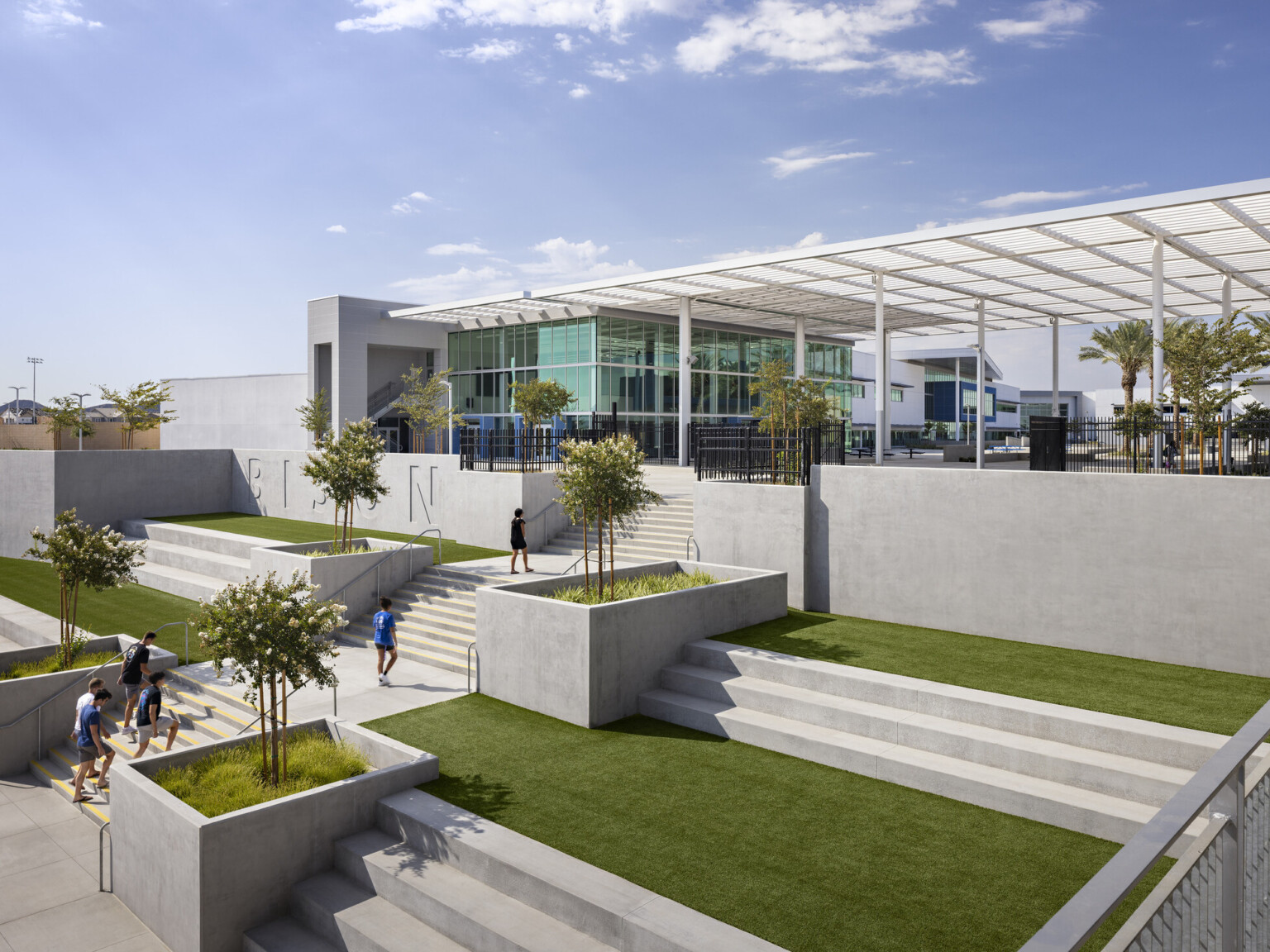 Concrete stairs lined with trees and learning steps with grass at landings, white canopy over courtyard outside glass facade school