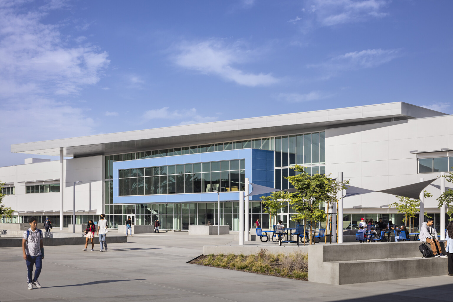 Front entrance of high school with blue mounted hallway on glass face. White angular canopy from roof, white wrap facade