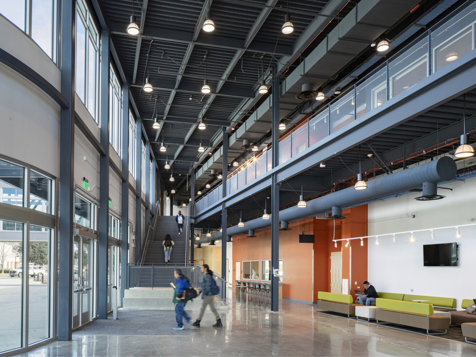 Double height hallway with large windows to courtyard. Industrial ceiling, exposed duct under upper level balcony walkway