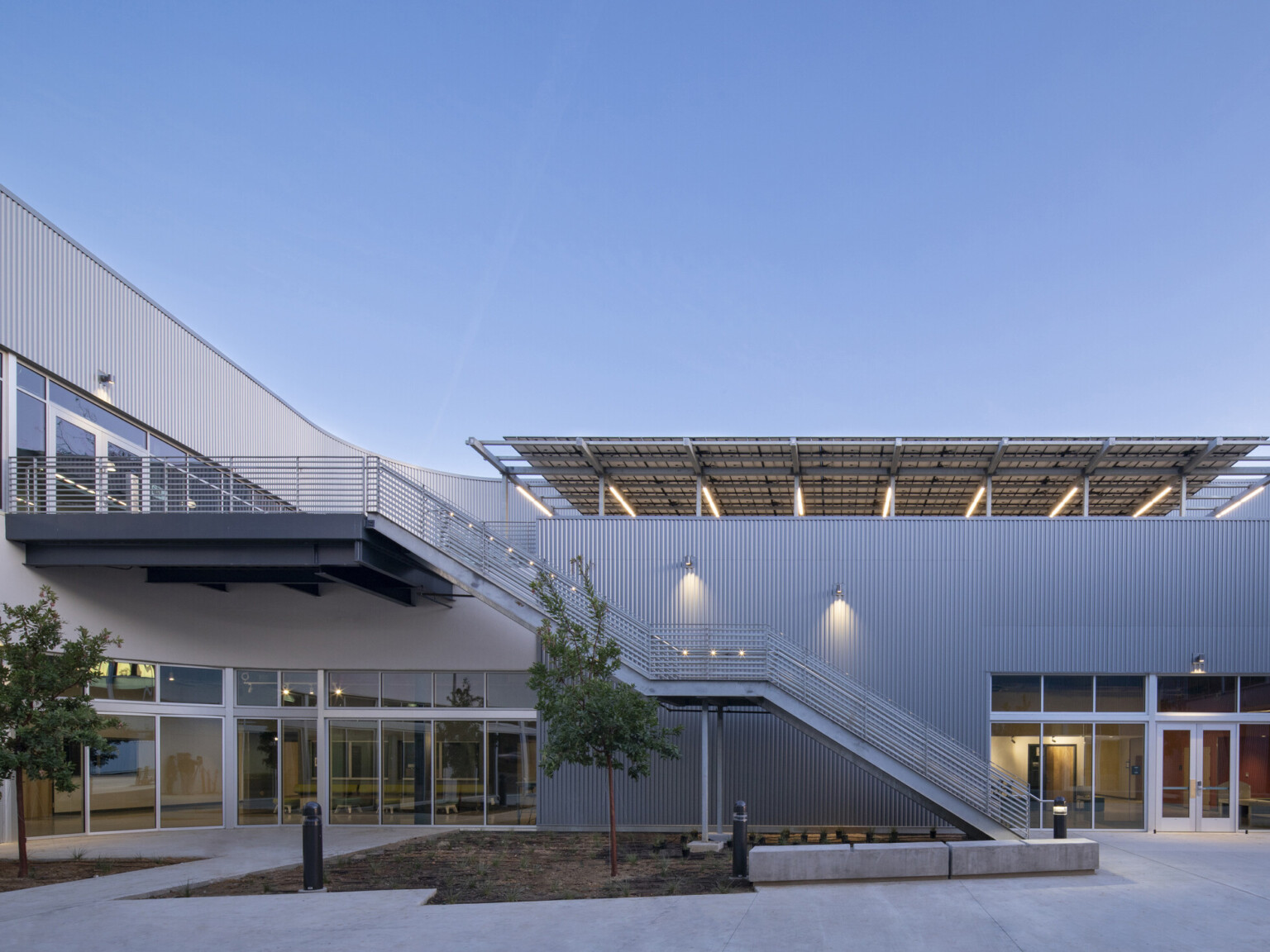 Exterior of 2 story arts complex with textural silver facade, large windows. Stairs to covered patio over single story, right