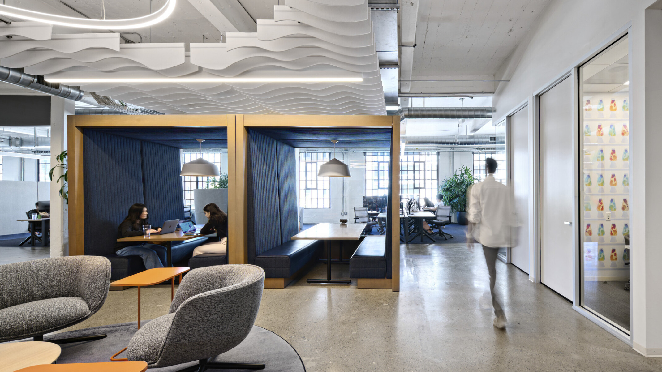 Office common space with grey armchairs and orange tables. 2 wood booths with bench seating at center enclosed front, back, above
