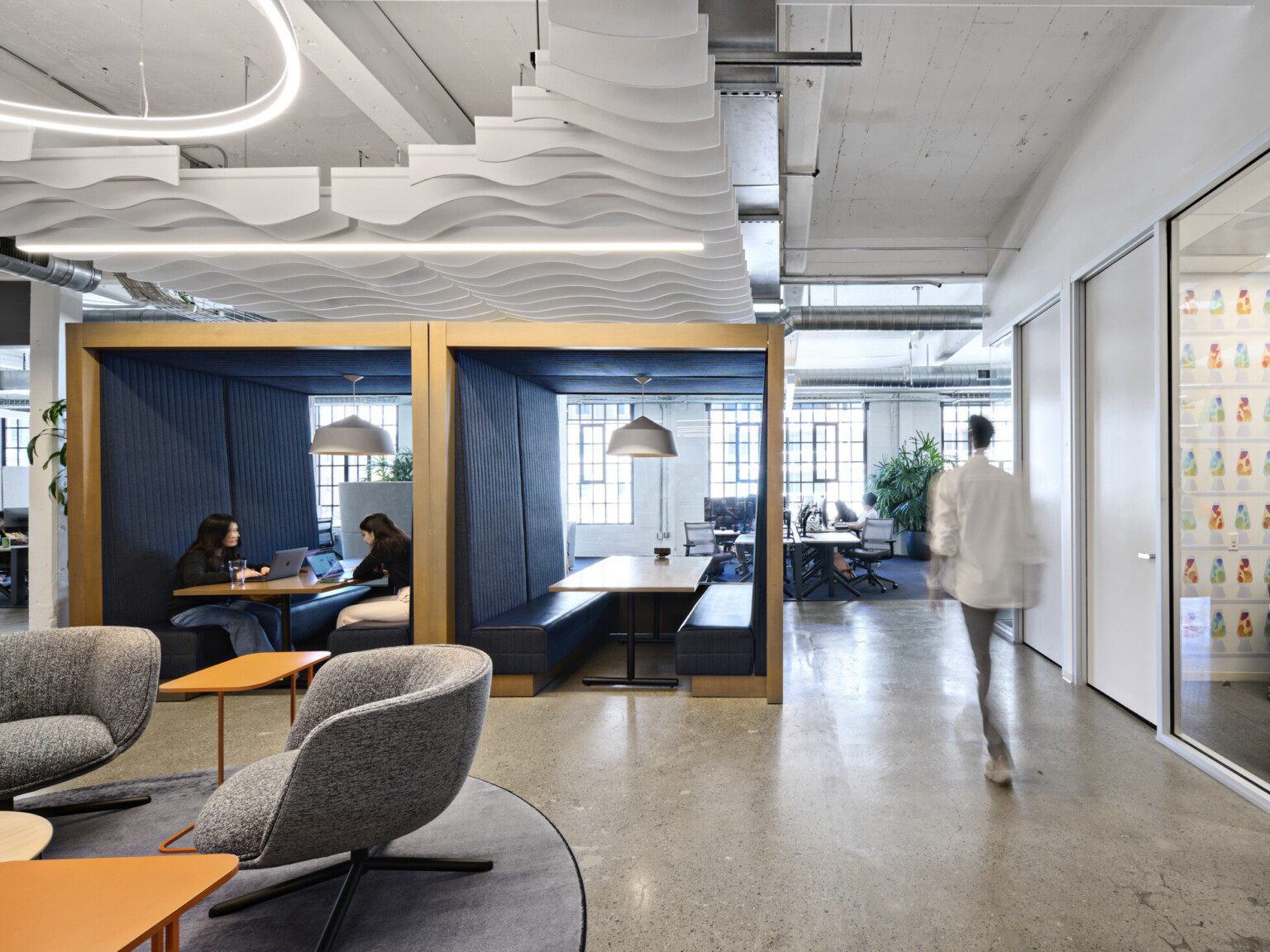 Office common space with grey armchairs and orange tables. 2 wood booths with bench seating at center enclosed front, back, above