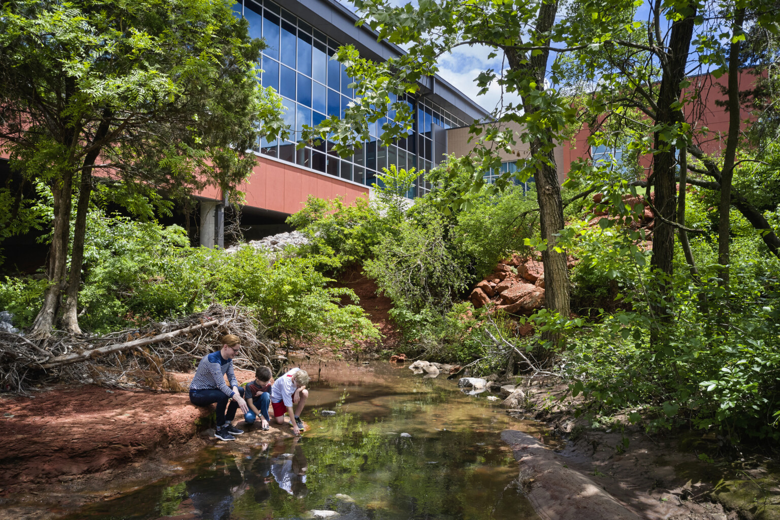 Two young students participating in outdoor learning near modern structure with floor-to-ceiling windows, lush landscaping and a reflecting pond