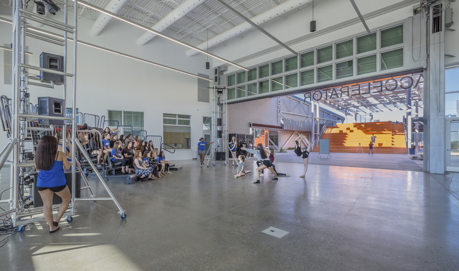 A white room, with the wall opened facing orange stairs, has portable bleachers and equipment