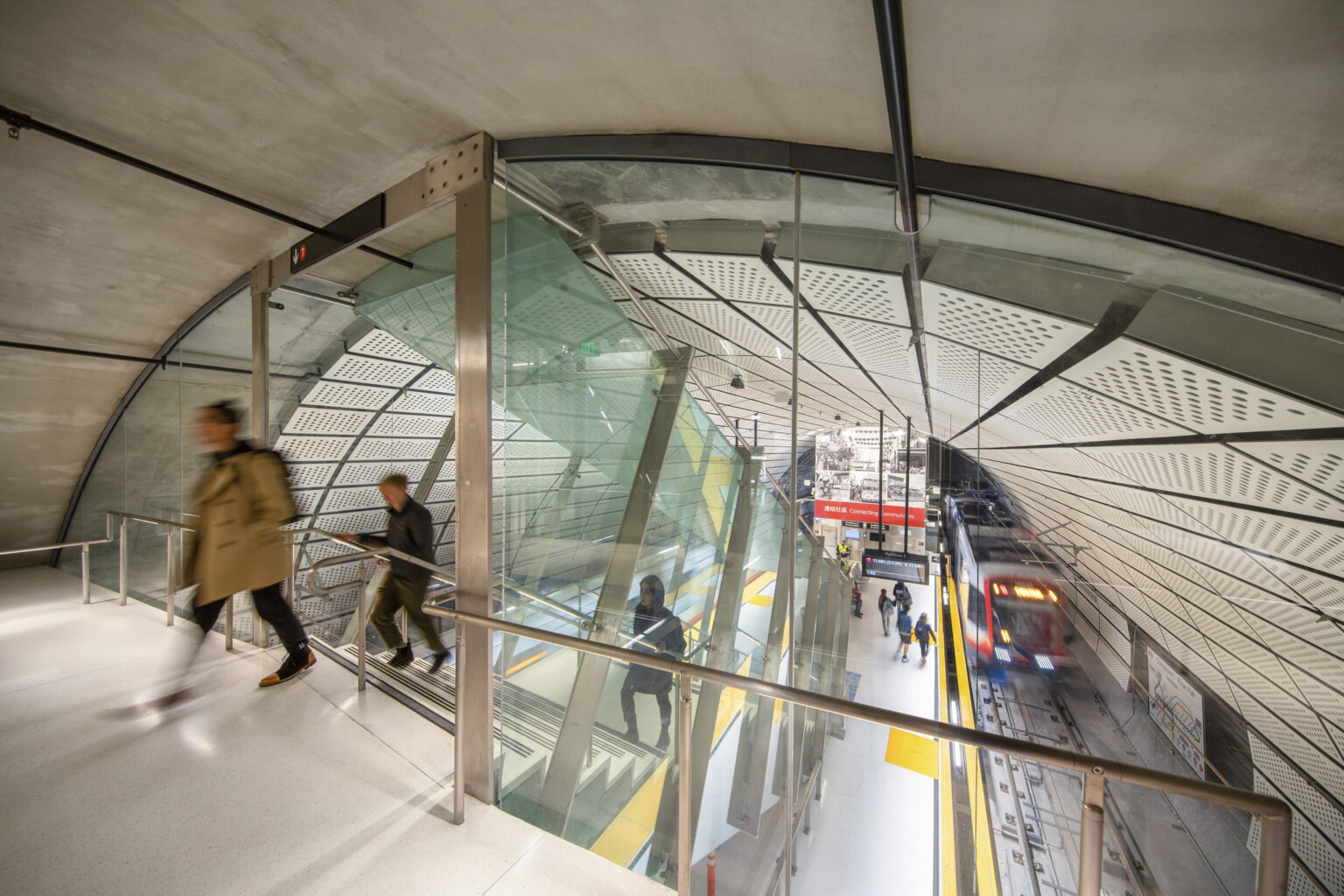 Rounded room with escalator to station below between 2 train tracks. White panel ceiling. Glass wall dividing tunnel