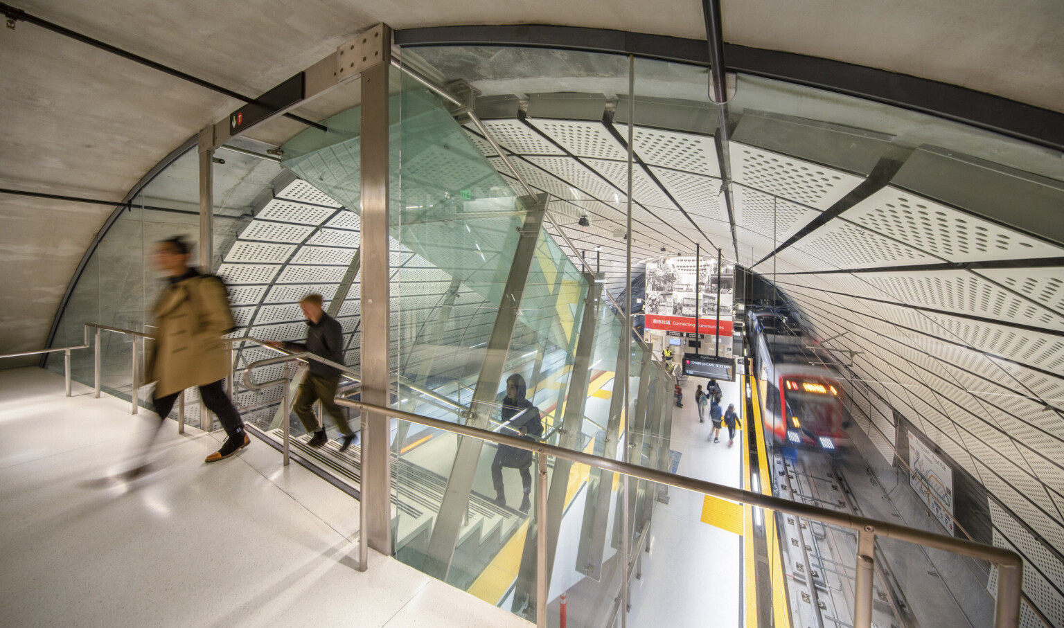 Rounded room with escalator to station below between 2 train tracks. White panel ceiling. Glass wall dividing tunnel