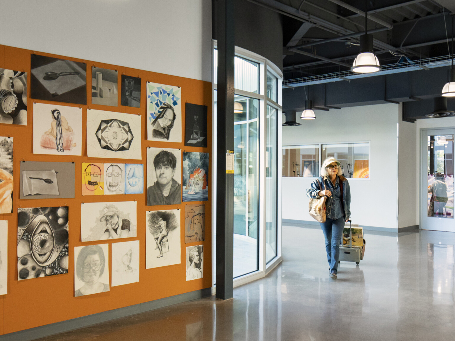 Interior rounded hallway with orange accent wall with student art work hanging on it, floor to ceiling window beyond