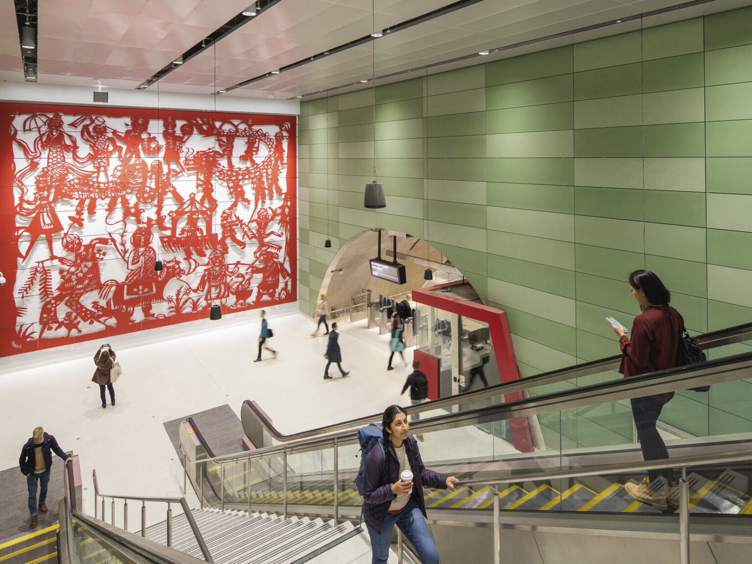 Concrete block walls with raised red sculptural mural on left. Right, arched hall with gates and ticket booth by escalators