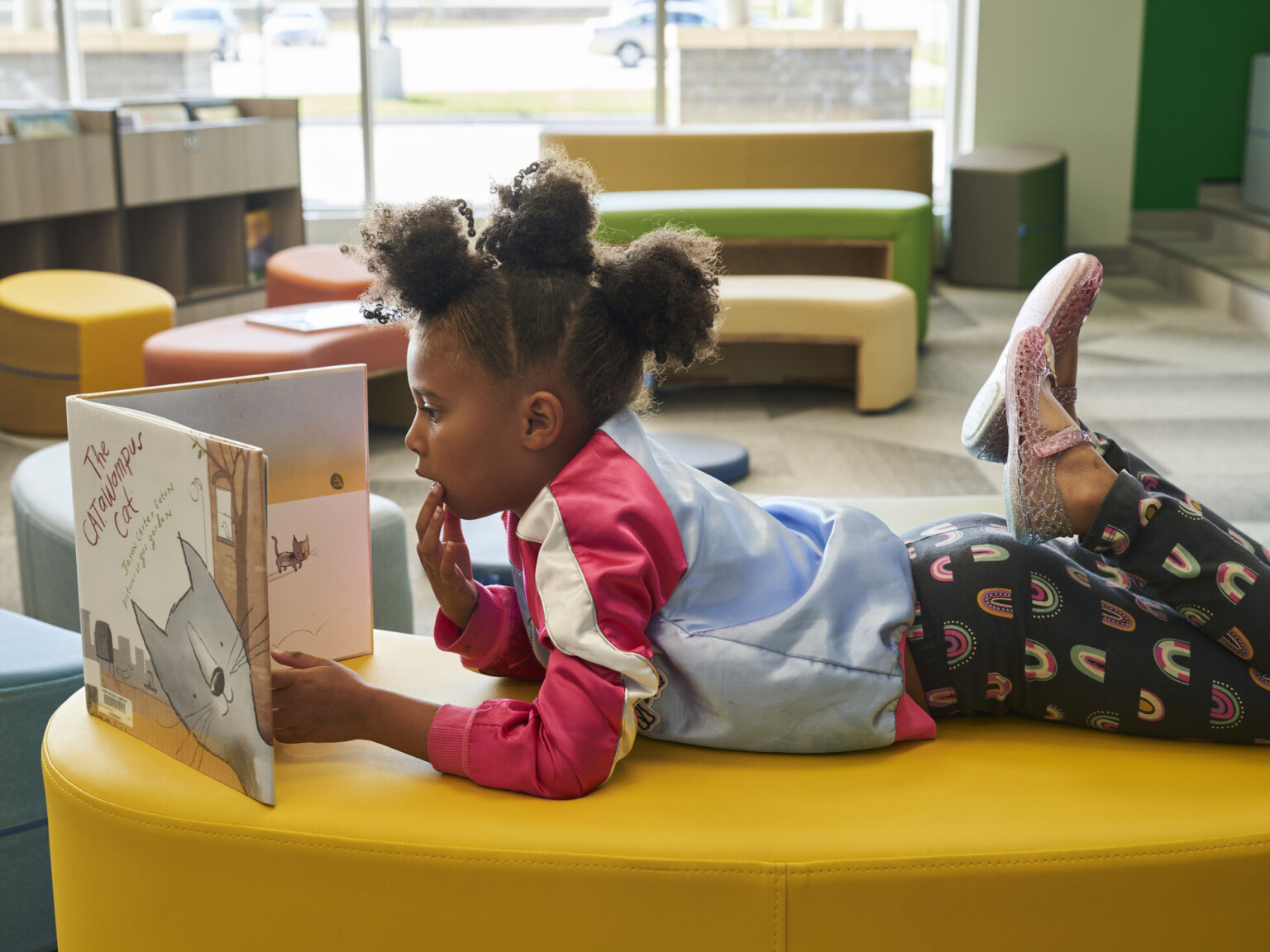 Grade school student relaxing on a comfy bright yellow cushion while reading a book in a day-lit sun-filled room