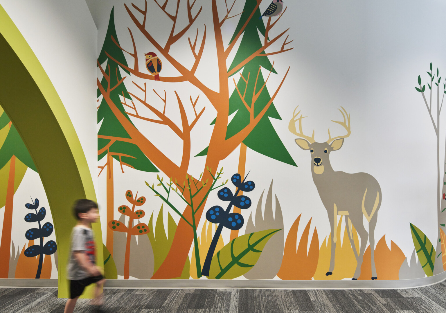 Child walks by colorful mural of cartoon forest and animals in carpeted school hallway, mixed trees, deer, owl, racoon