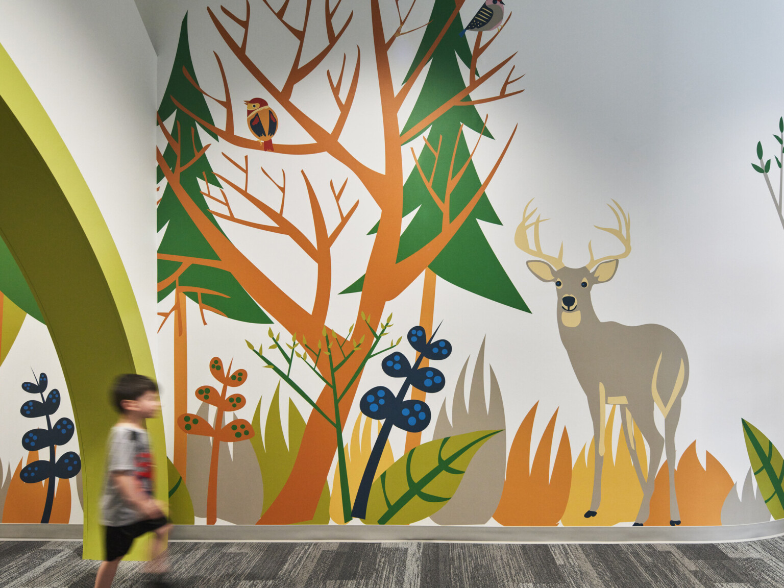 Child walks by colorful mural of cartoon forest and animals in carpeted school hallway, mixed trees, deer, owl, racoon