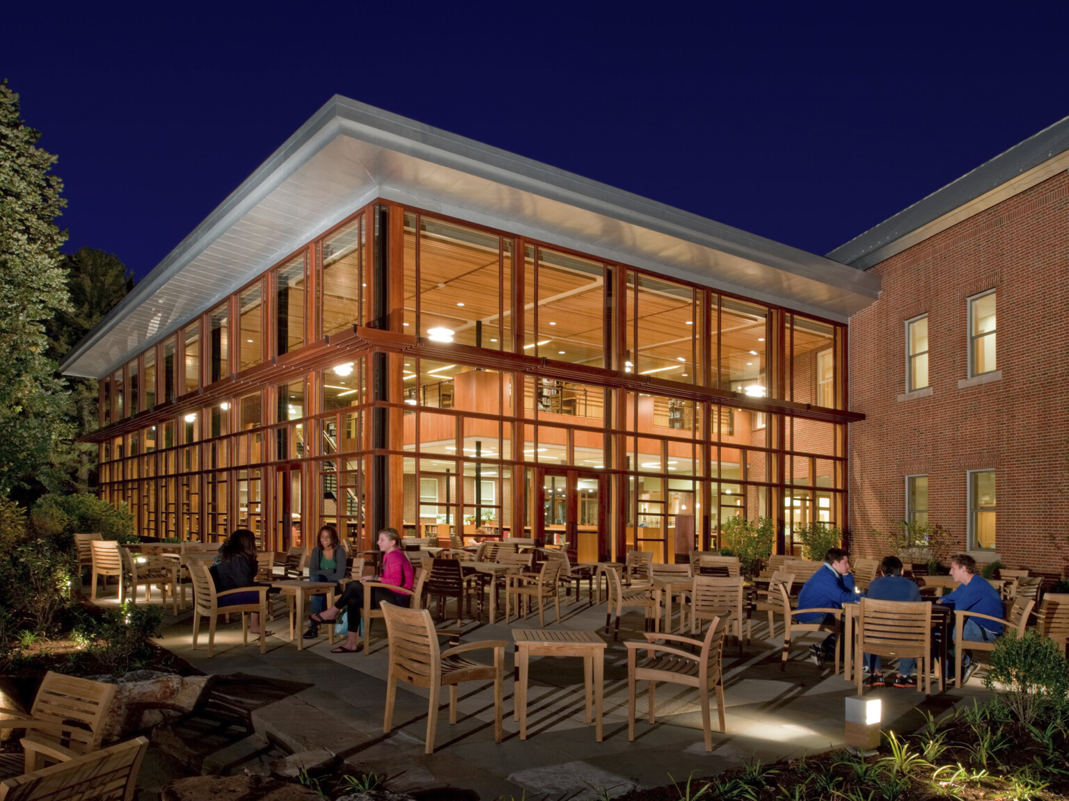 Wood framed double height glass facade library addition to brick building illuminated at night, seen from large patio seating area