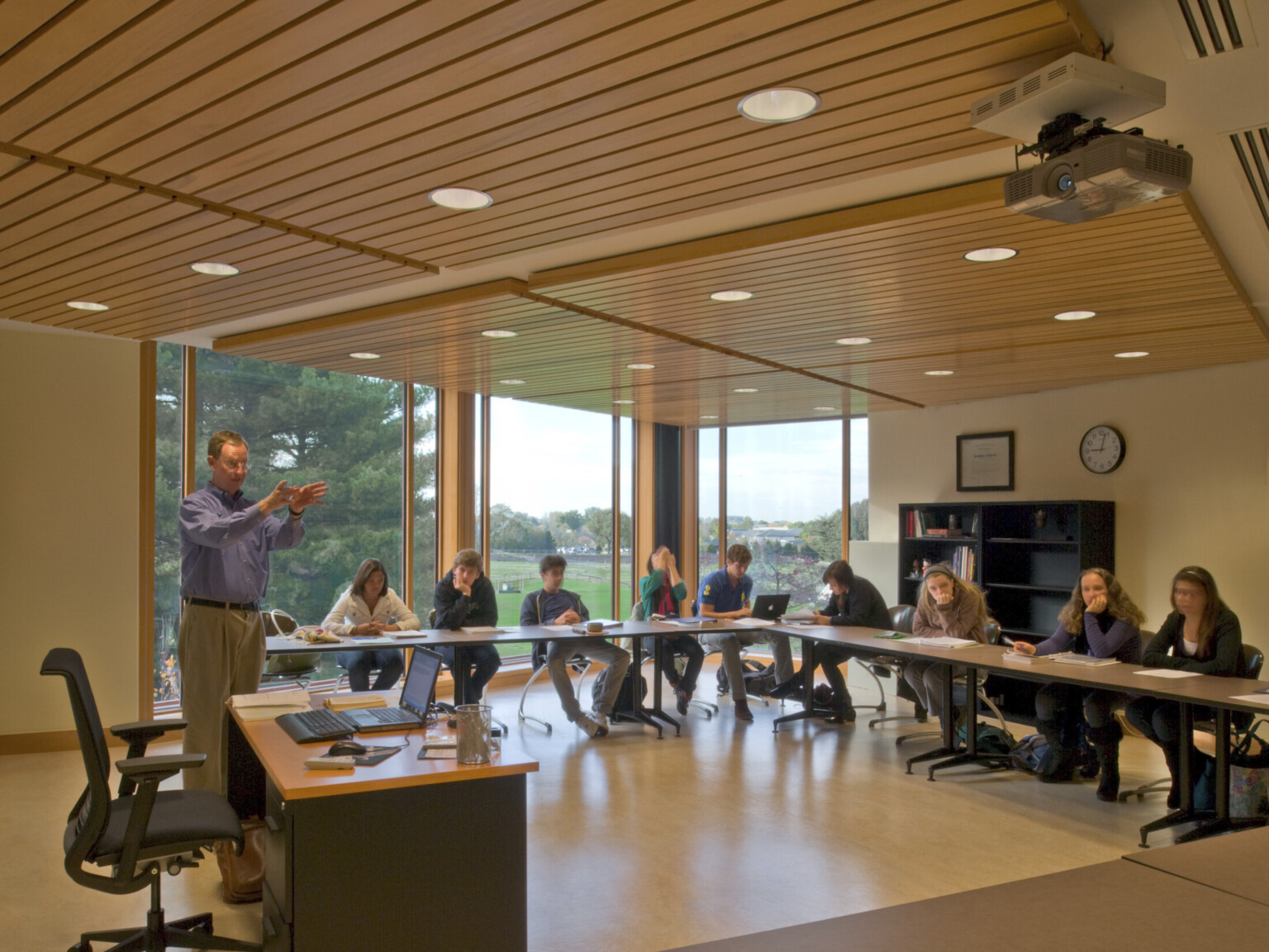Conference room learning space with floor to ceiling windows, students sit at long tables angled toward desk and projector