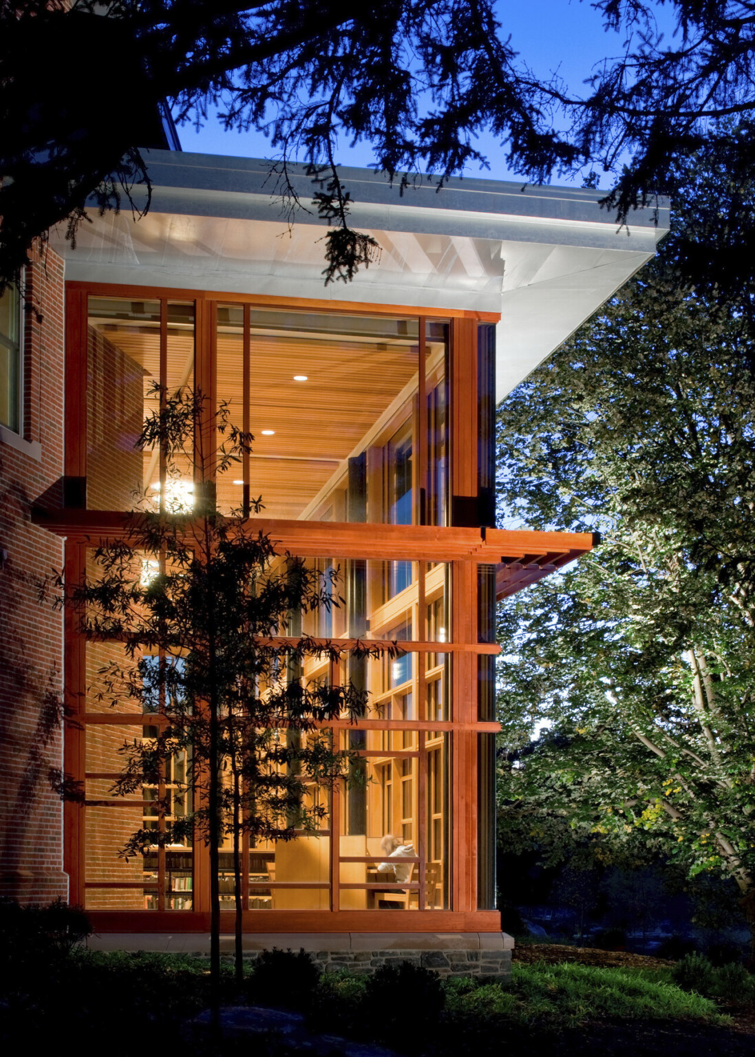 Exterior view of corner space, double height space illuminated at night, surrounded by trees