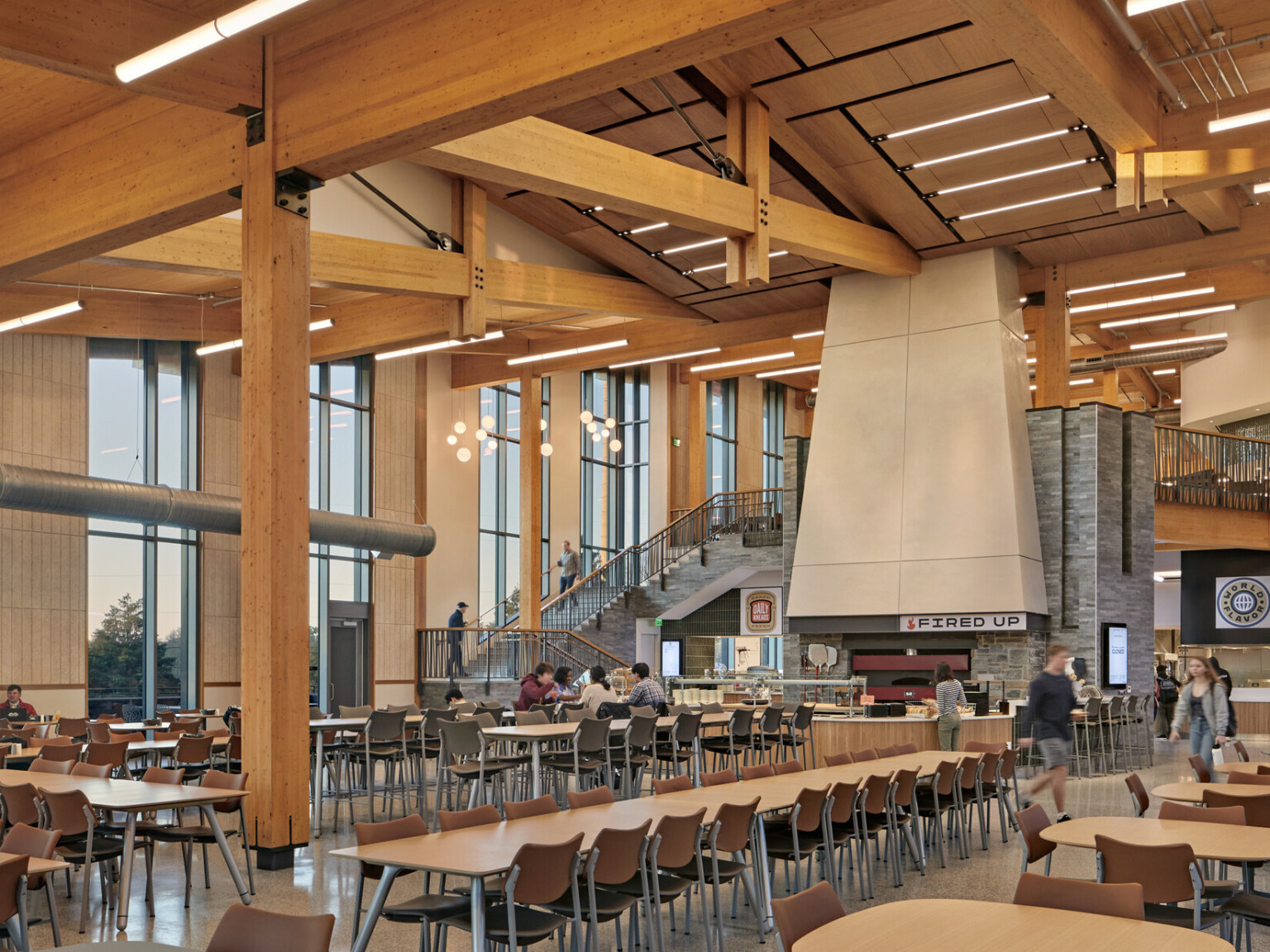 large two-story glass dining hall with exposed timber beams and posts, stone wrapped stair leading to second floor seating area