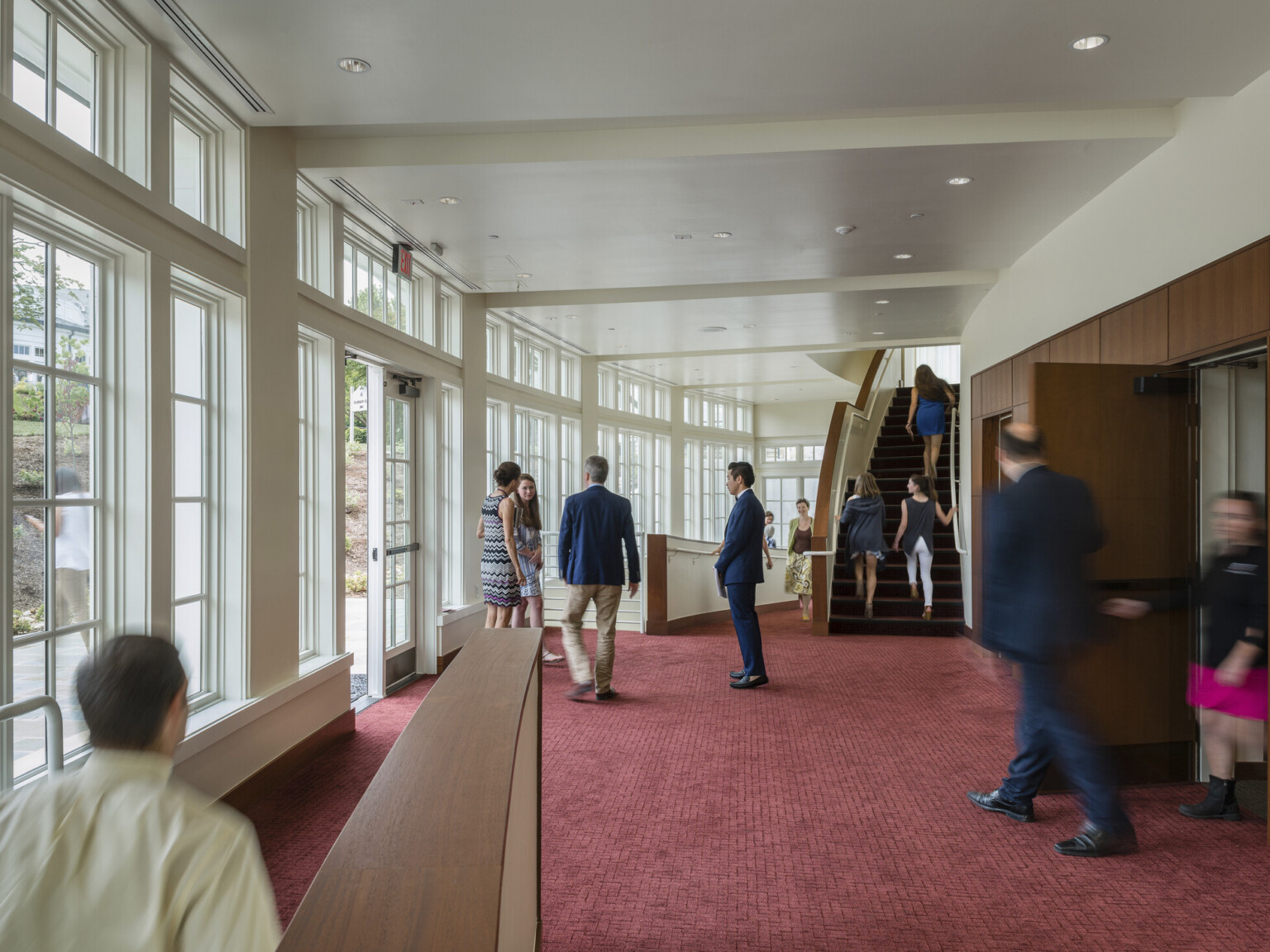 Auditorium theater lobby with framed floor to ceiling windows on rounded white left wall, red carpet, stairs to balcony seats