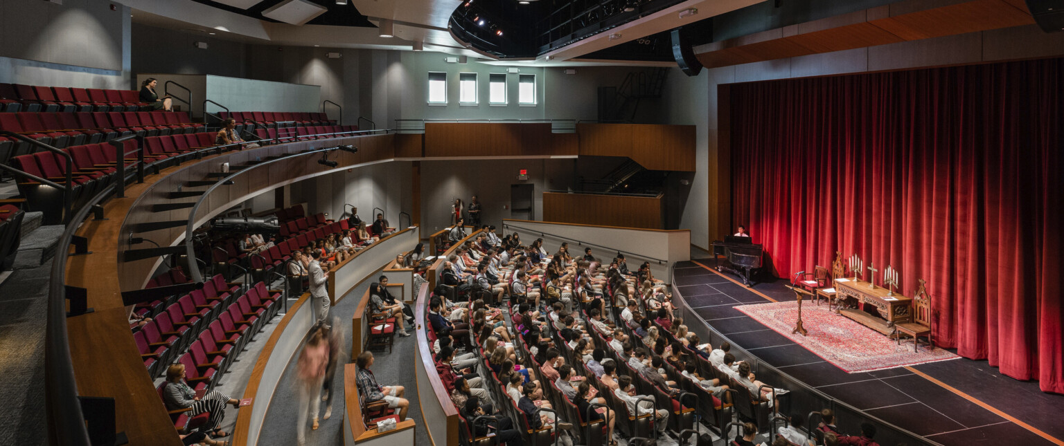 Double height auditorium theater with rounded rows of seating facing stage with red curtains, brown acoustic panels above